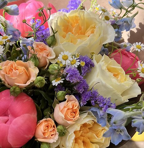 A close-up photograph of a mixed floral bouquet containing peach roses, cream lisianthus, purple statice, and white daisies.