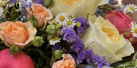 A close-up photograph of a mixed floral bouquet containing peach roses, cream lisianthus, purple statice, and white daisies.