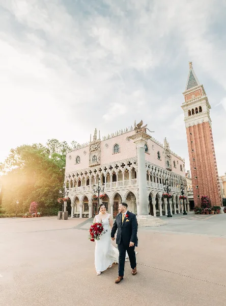 A bride and groom walking hand-in-hand in front of a pink and white Venetian-style building with a tall brick tower.