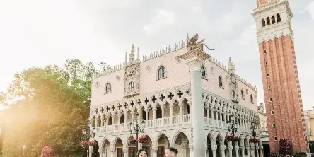 A bride and groom walking hand-in-hand in front of a pink and white Venetian-style building with a tall brick tower.