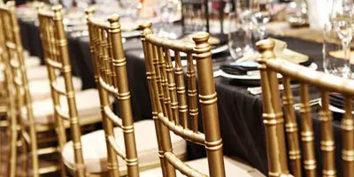 Rows of gold chiavari chairs set up at a dining table with tableware and glassware.