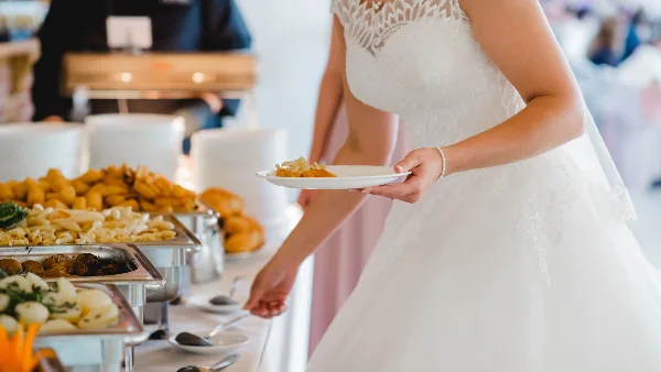 A bride in a white lace wedding dress holding a plate of food while standing in front of a catering buffet with various dishes.