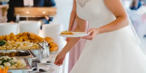 A bride in a white lace wedding dress holding a plate of food while standing in front of a catering buffet with various dishes.
