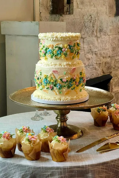 A two-tiered white wedding cake decorated with colorful buttercream flowers on a gold stand, surrounded by matching cupcakes.
