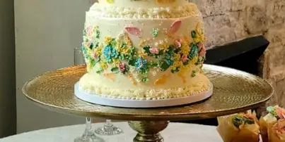 A two-tiered white wedding cake decorated with colorful buttercream flowers on a gold stand, surrounded by matching cupcakes.