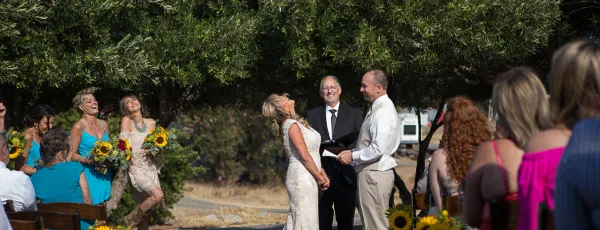 A wedding officiant in a black suit stands between a bride and groom during an outdoor ceremony with seated guests.