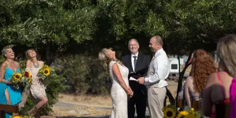 A wedding officiant in a black suit stands between a bride and groom during an outdoor ceremony with seated guests.