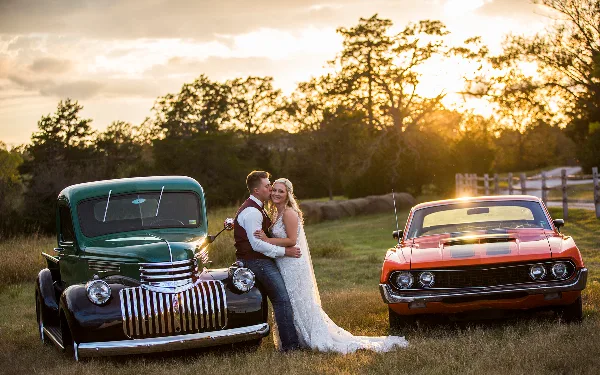 A bride and groom posing together in a field between a vintage green truck and a red muscle car during a sunset.
