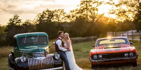 A bride and groom posing together in a field between a vintage green truck and a red muscle car during a sunset.