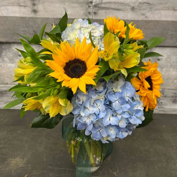 A floral arrangement containing sunflowers, blue hydrangeas, and yellow alstroemeria in a glass vase.