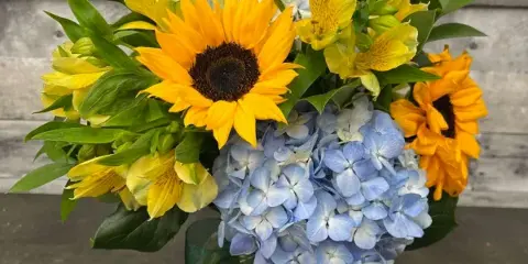 A floral arrangement containing sunflowers, blue hydrangeas, and yellow alstroemeria in a glass vase.