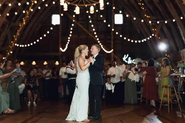 A bride and groom dancing together at a wedding reception in a barn venue with string lights.