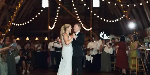 A bride and groom dancing together at a wedding reception in a barn venue with string lights.