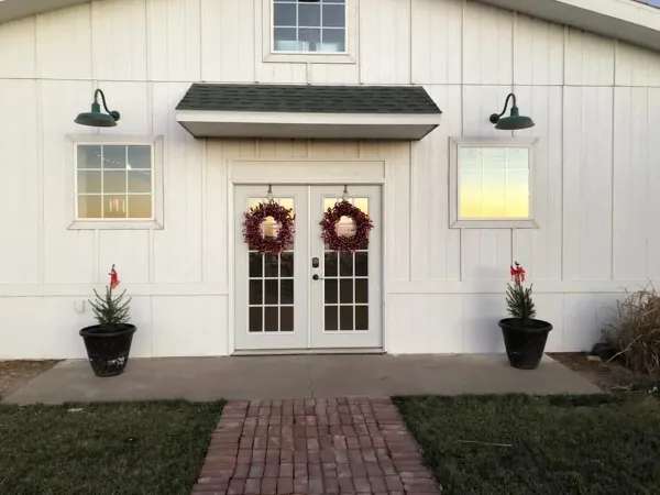 Exterior view of a white barn venue with double glass doors, brick walkway, and holiday wreaths.