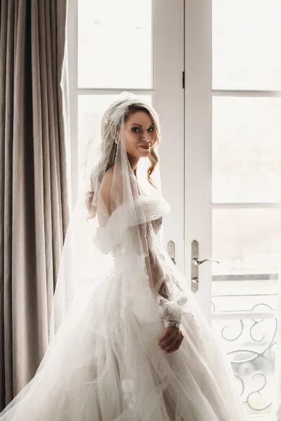 A bride with styled hair and makeup wearing a white wedding dress and veil, standing near a window.