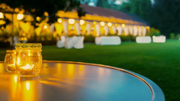 An outdoor event setup featuring a large white tent, white chair covers, and a table with lit candles in the foreground.