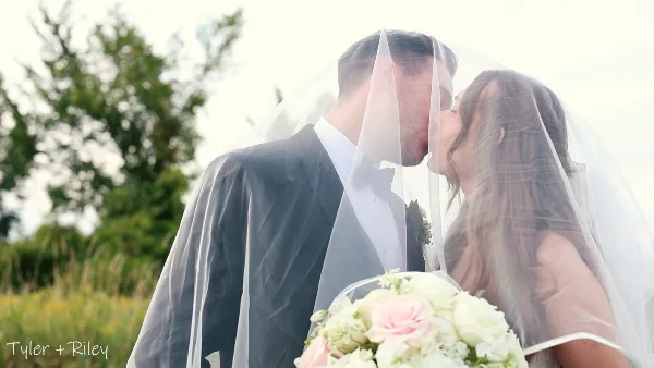 A cinematic wedding video still showing a bride and groom sharing a kiss under a veil in an outdoor setting.