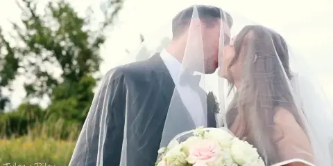 A cinematic wedding video still showing a bride and groom sharing a kiss under a veil in an outdoor setting.