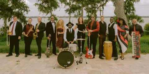 A group of musicians and vocalists posing outdoors with their instruments, including a drum kit, electric guitar, saxophone, and accordion.