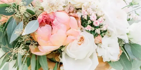 A close-up photograph of a wedding bouquet with white and peach roses, peonies, and greenery tied with a ribbon.