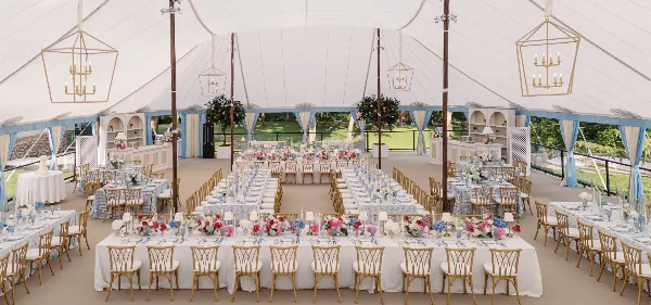 Luxury wedding reception tent interior featuring long banquet tables, floral centerpieces, gold chairs, and geometric chandeliers.