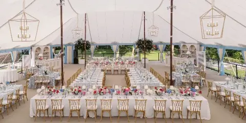 Luxury wedding reception tent interior featuring long banquet tables, floral centerpieces, gold chairs, and geometric chandeliers.
