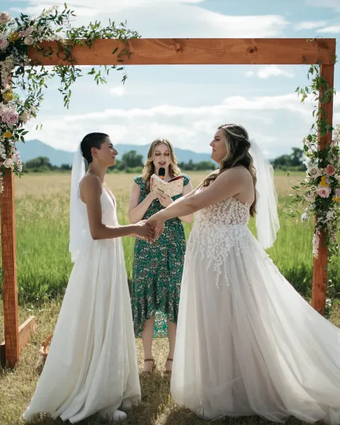 Two brides exchanging vows under a wooden arch decorated with floral arrangements in a grassy field.