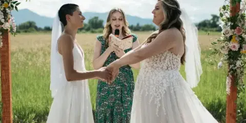 Two brides exchanging vows under a wooden arch decorated with floral arrangements in a grassy field.