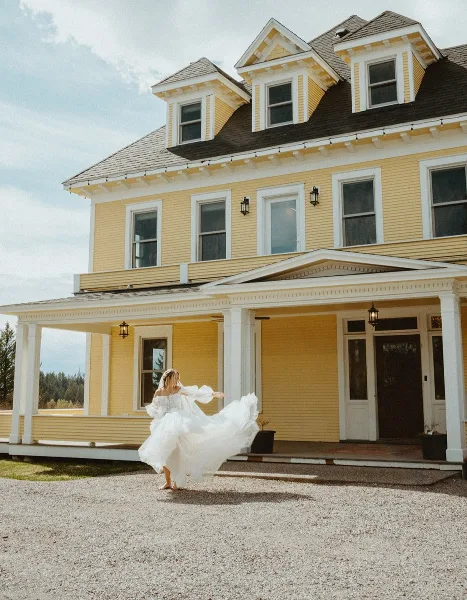 A bride in a white wedding dress twirling in front of a large, historic yellow mansion with white trim and a porch.