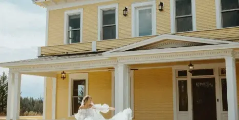 A bride in a white wedding dress twirling in front of a large, historic yellow mansion with white trim and a porch.