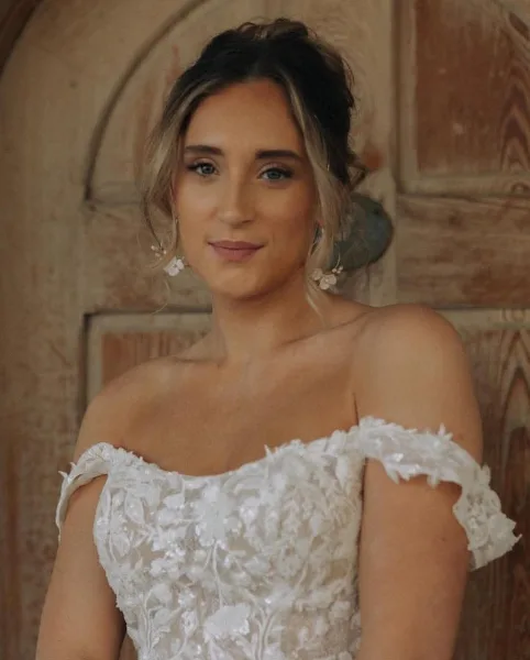 A bride wearing an off-the-shoulder white lace wedding dress with a soft updo hairstyle and natural makeup.