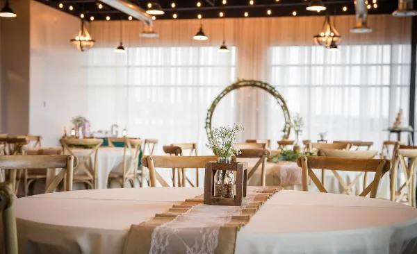 A wedding reception hall with round tables, wooden chairs, and a floral arch in the background.