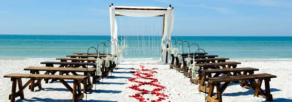 A beach wedding ceremony setup featuring a bamboo arch, white drapery, and wooden benches on white sand.