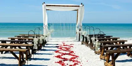 A beach wedding ceremony setup featuring a bamboo arch, white drapery, and wooden benches on white sand.