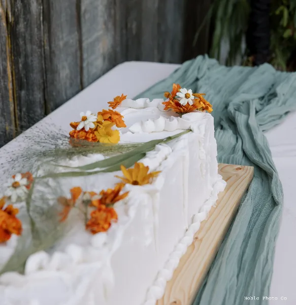 A white frosted wedding cake decorated with orange and white flowers on a wooden board.