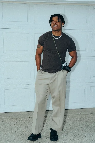 A person standing in front of a white garage door wearing a dark t-shirt and light pants, with a camera hanging from a strap.