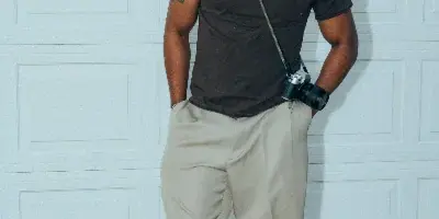 A person standing in front of a white garage door wearing a dark t-shirt and light pants, with a camera hanging from a strap.