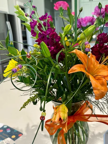 A vibrant floral arrangement with orange lilies, purple stock, and greenery in a glass vase with an orange ribbon.