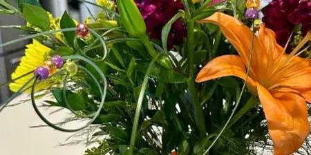 A vibrant floral arrangement with orange lilies, purple stock, and greenery in a glass vase with an orange ribbon.