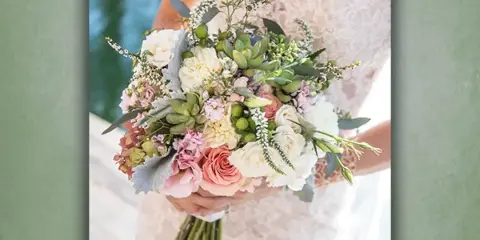 A bride in a lace dress holding a mixed bouquet of white and pink flowers with greenery.