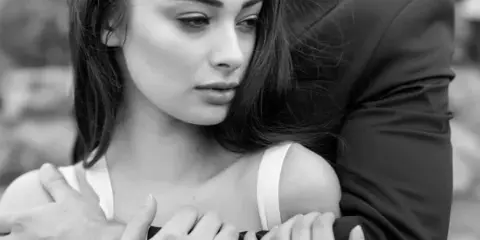 Black-and-white close-up of a bride's face showing styled hair and bridal makeup, leaning against a groom in a suit.