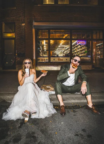 Bride in a white gown and groom in a green suit sitting on a curb at night eating pizza and drinking from a cup in front of a storefront.