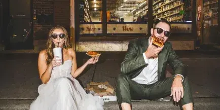 Bride in a white gown and groom in a green suit sitting on a curb at night eating pizza and drinking from a cup in front of a storefront.