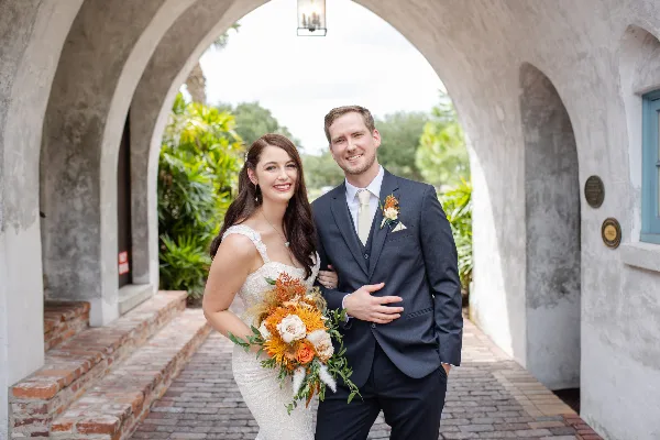 A bride and groom standing together, with the bride holding a bouquet of orange and white flowers and the groom wearing a matching boutonniere.