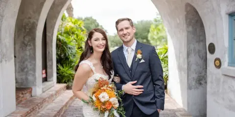 A bride and groom standing together, with the bride holding a bouquet of orange and white flowers and the groom wearing a matching boutonniere.