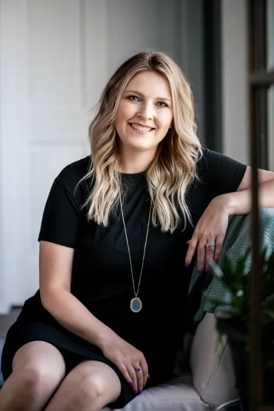 Professional portrait of a smiling woman with blonde hair wearing a black dress and a long pendant necklace, seated in a studio setting.