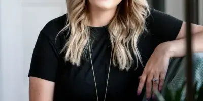 Professional portrait of a smiling woman with blonde hair wearing a black dress and a long pendant necklace, seated in a studio setting.
