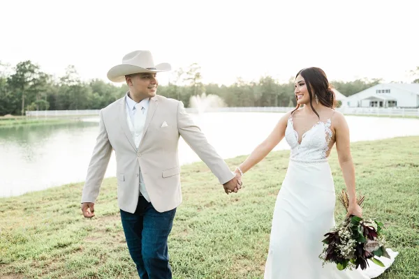 A bride and groom holding hands and walking on the grass near a pond at a countryside event venue.