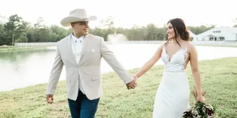 A bride and groom holding hands and walking on the grass near a pond at a countryside event venue.