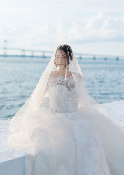 A bride in a white lace wedding dress and veil sitting by the water.